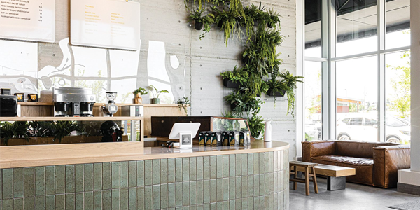 A modern, minimalistic café interior featuring a tiled coffee bar with wood accents, a living plant wall, and floor to ceiling windows. A leather couch and small table sits in the corner, creating a bright, inviting space.