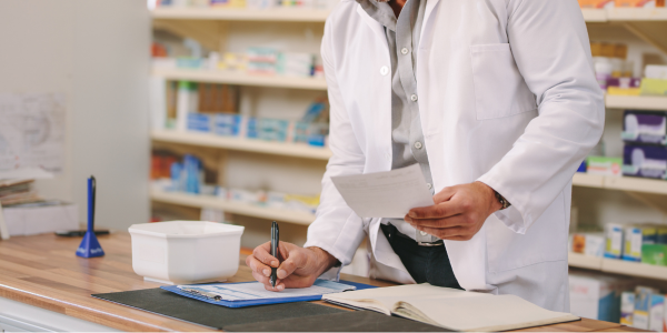 Image of a male pharmacist writing on paper clipped to a blue clipboard on a counter, with shelves full of medication behind him.