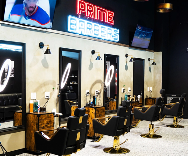 A modern barbershop interior with sleek black and gold barber chairs, large mirrors, and ring lights at each station. A neon sign reading "Prime Barbers" is mounted on the back wall above TVs showing sports.