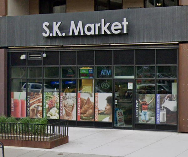 Street view of S.K. Market storefront in Calgary, featuring large window decals advertising slushies, hot dogs, ice cream, samosas, cold drinks, lottery tickets, and dairy products, with a prominent sign above the entrance.