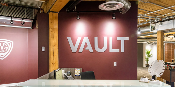 Interior view of Vault Group’s office featuring a modern industrial design with exposed wood beams and ductwork. A maroon wall displays the silver VAULT logo behind a reception desk.