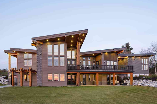 A modern, custom-built home with large floor-to-ceiling windows, sloped rooflines, and a mix of wood and stone exterior. This home features warm exterior lighting and sits on a spacious, well-kept lawn.