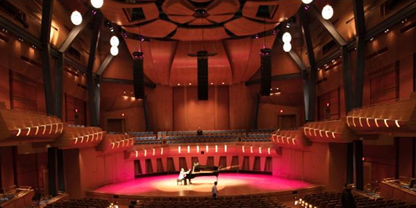 A spacious and modern concert hall with wood paneling and dramatic lighting. A grand piano sits centre stage, with a person on stage under focused lighting, highlighting the hall's acoustic and visual design feature.