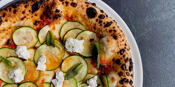 Close-up of a wood-fired pizza topped with thinly sliced zucchini, dollops of ricotta cheese, fresh mint leaves, and melted cheese on a charred, crispy crust. Served on a white plate against a dark textured surface.