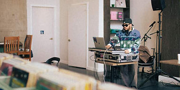 A DJ performing on a turntable setup inside Semantics Cafe, with vinyl records in the foreground, wooden chairs, a mic stand, and bookshelves in the background.