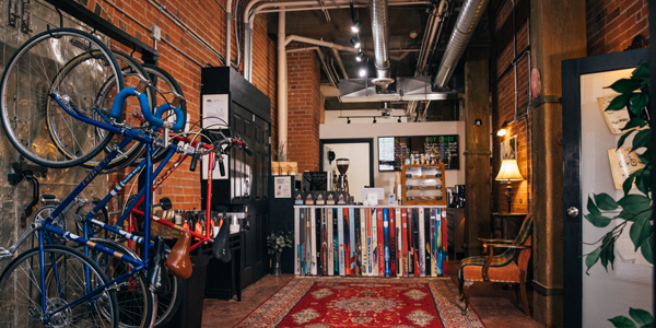 Interior of Velet Bike Ski Cafe, a cozy bike and ski repair shop with a vintage-inspired coffee counter made from repurposed skis. The space features hanging bicycles, exposed brick walls, a Persian rug, and warm lighting, blending workshop and cafe charm.