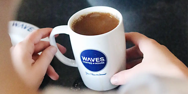 Close-up of a person holding a white Waves Coffee House mug filled with coffee. The mug features the blue Waves Coffee House logo and the tagline “Your Place to Connect.”